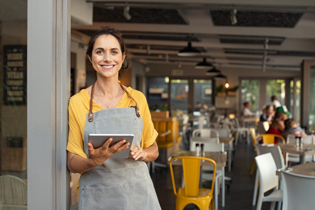 Portrait of happy woman standing at doorway of her franchise store holding digital tablet. Successful franchise business owner in casual clothing and grey apron standing at entrance and looking at camera.